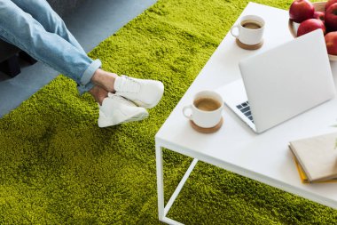 kahve fincanı, dizüstü bilgisayar, kitap ve evde elma içeren tablo yakınındaki kanepede oturan adam kırpılmış atış