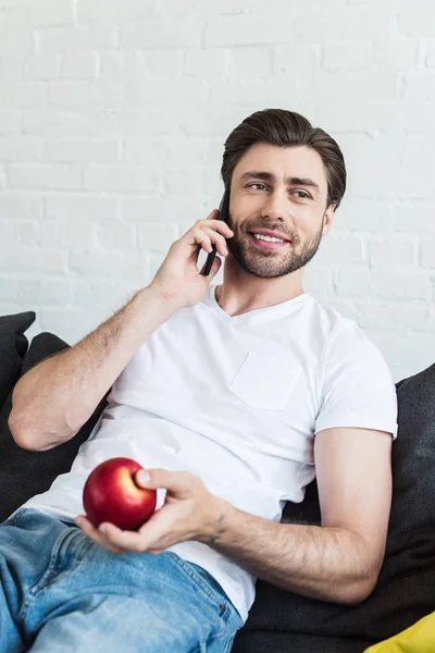 Hombre Sonriente Hablando Teléfono Inteligente Sosteniendo Manzana Mano Casa — Foto de Stock