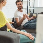 Young smiling couple with coffee cups talking and sitting on sofa at home