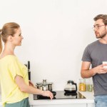 Vista laterale della donna sorridente che cucina sul fornello e parla con il fidanzato con una tazza di caffè in cucina