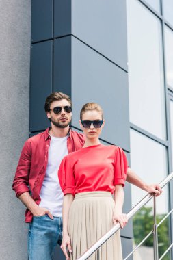 front view of stylish young couple of models in sunglasses standing on staircases  clipart