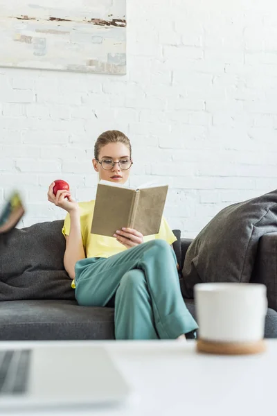 Ελκυστική Γυναίκα Γυαλιά Κρατώντας Apple Και Ανάγνωση Βιβλίων Στο Σπίτι — Φωτογραφία Αρχείου