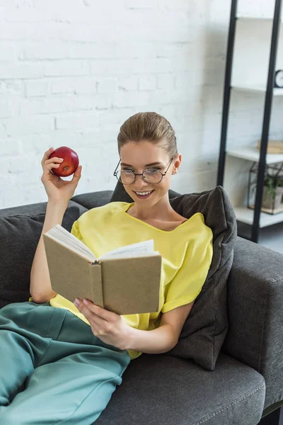 Joven Mujer Sonriente Gafas Con Manzana Libro Lectura Sofá Casa — Foto de Stock