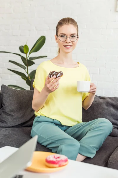 Jovem Mulher Óculos Beber Café Comer Donut Sofá Casa — Fotografia de Stock