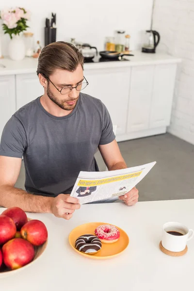 リンゴ ドーナツとコーヒー カップとテーブルで新聞を読む眼鏡の若い男  — 無料ストックフォト