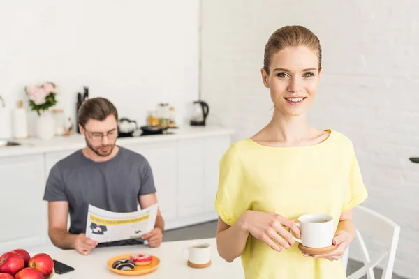 コーヒー カップとテーブルに座って キッチンで新聞を読んで彼氏を保持している魅力的な笑顔の女性  — 無料ストックフォト