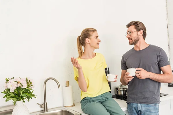 愉快的年轻夫妇与咖啡杯子谈话在厨房 — 图库照片