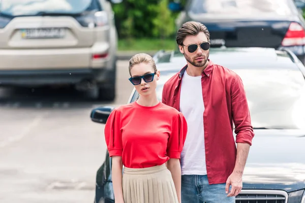 stylish young couple of models in sunglasses posing near car