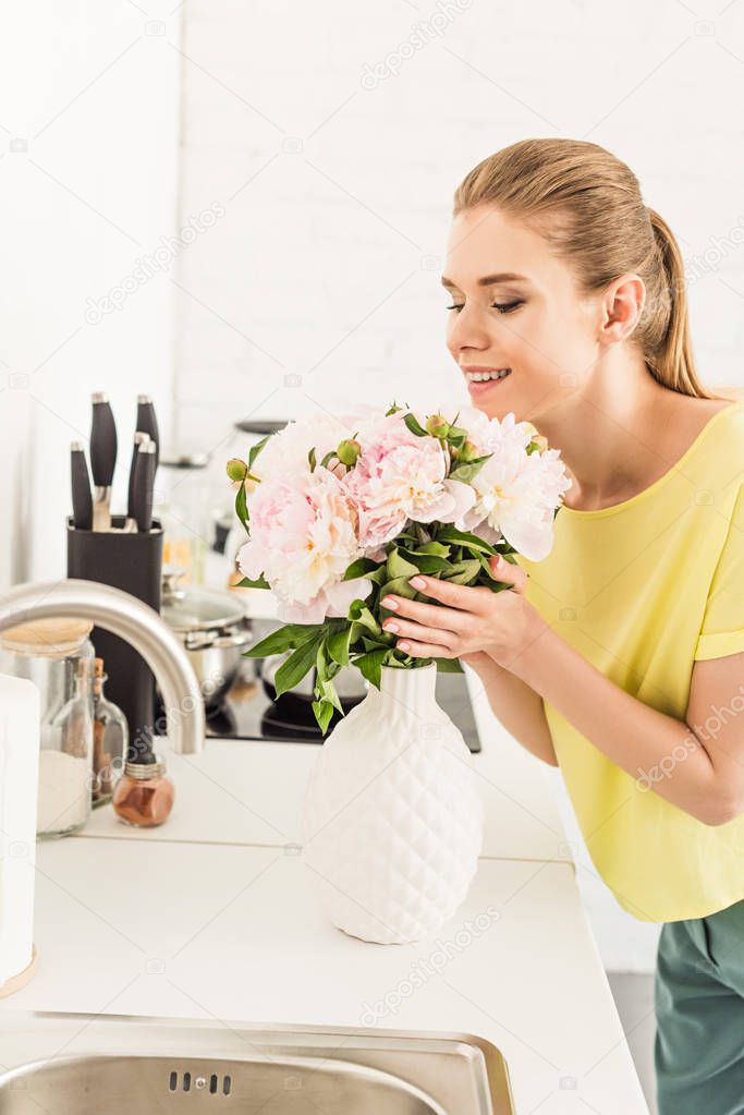 young smiling woman sniffing bouquet of peonies at kitchen