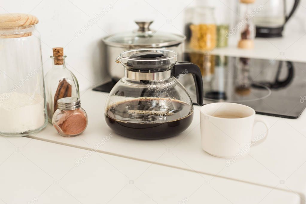 close up view of coffee maker, cup and glass jars near stove on tabletop at kitchen