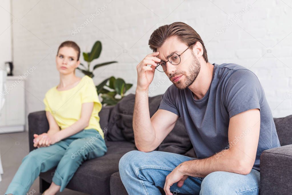 upset young man in eyeglasses and girlfriend sitting on sofa behind at home
