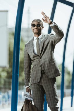 Young african american businessman waving to someone on train station clipart