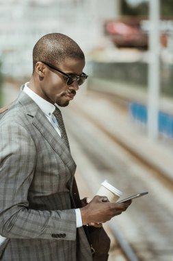 Genç Afro-Amerikalı işadamı ile tren bekliyorsun smartphone ve kahve Kupası