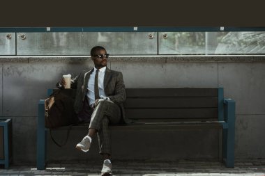 Stylish confident businessman with coffee cup sitting on train station bench clipart
