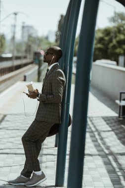 Müzik ve kahve fincanı tren istasyonu üzerinde tutan şık emin iş adamı