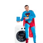 Super héros masculin souriant assis sur la machine à laver et pointant du doigt la poudre de savon sur blanc