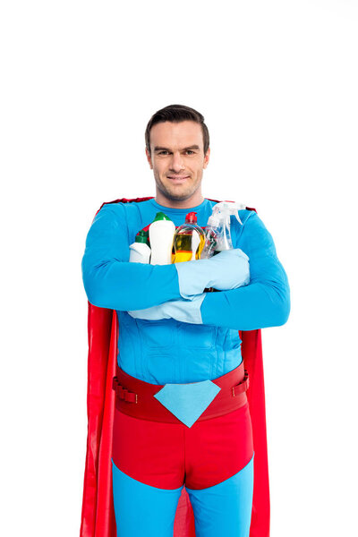 superman in rubber gloves holding cleaning items and smiling at camera isolated on white