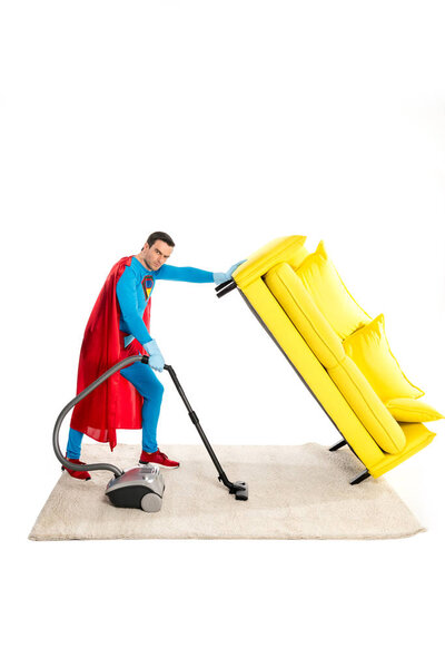 male superhero holding couch while cleaning carpet with vacuum cleaner and looking at camera isolated on white