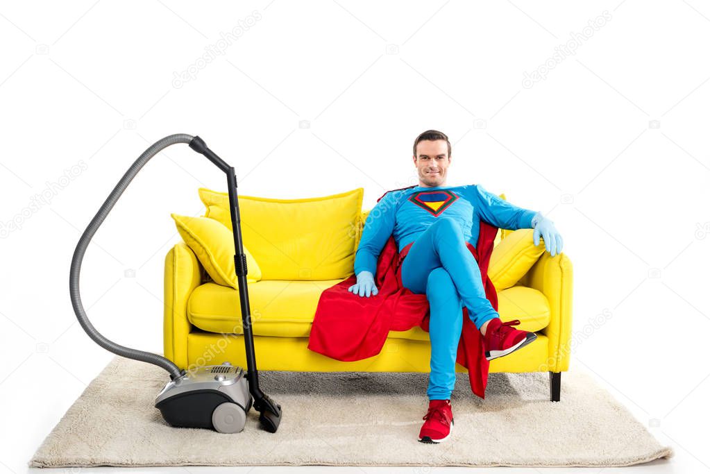 smiling male superhero in rubber gloves sitting on sofa near vacuum cleaner and looking at camera isolated on white