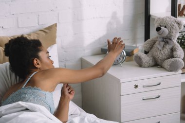 african american girl waking up and turning off alarm clock in bedroom clipart