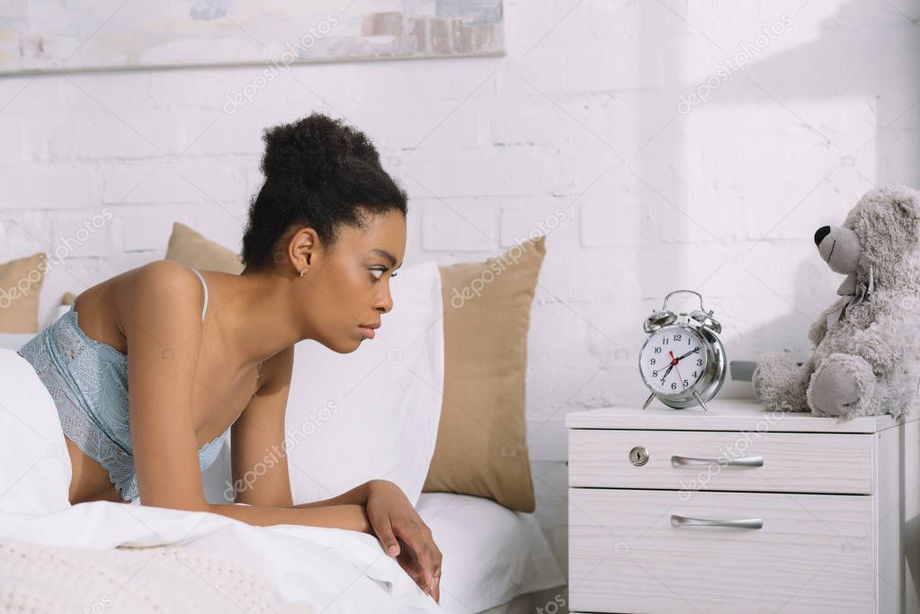 upset african american woman looking at alarm clock while lying in bed