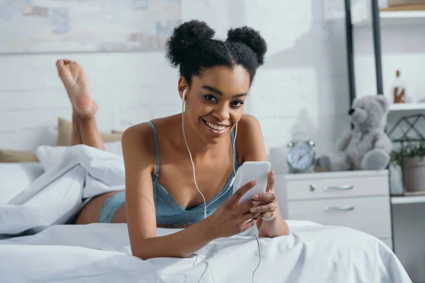 迷人的微笑非洲裔美国女孩在内衣听音乐与耳机和智能手机在床上 — 图库照片