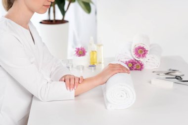 cropped shot of woman holding hands at table with towels, flowers, candles, aroma oil bottles and instruments for manicure in beauty salon clipart