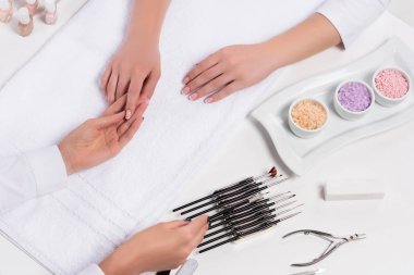 cropped shot of beautician doing manicure to woman at table with colorful sea salt, nail polishes, nail clippers, and tools for manicure in beauty salon  clipart