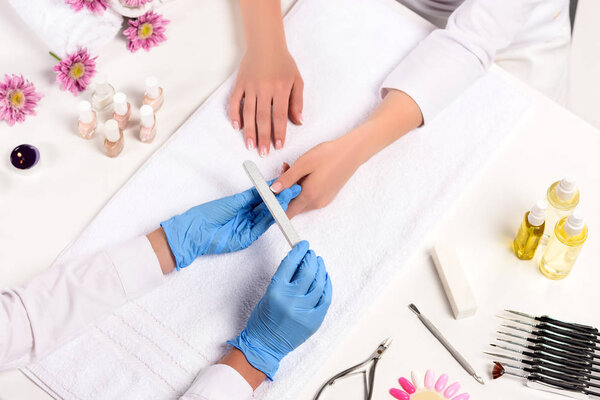 elevated view of beautician in latex gloves doing manicure to woman by nail file at table with flowers, towels, nail clippers, candles, nail polishes, aroma oil bottles and cuticle pusher  