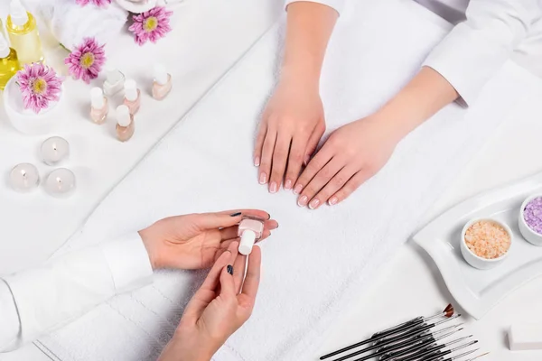 cropped image of manicurist showing nail polish to woman at table with candles, sea salt, flowers, aroma oil bottles, towels and tools for manicure