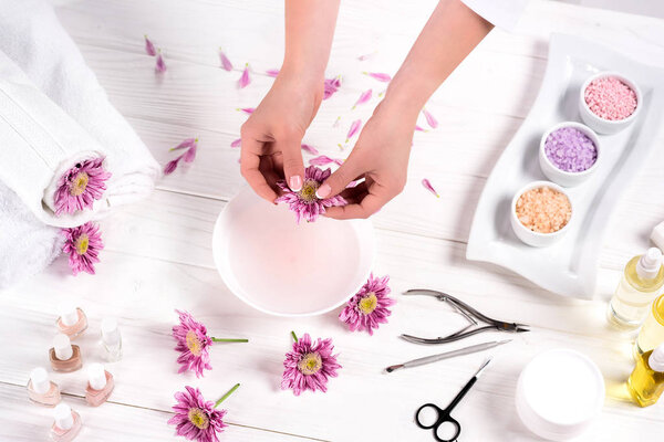 partial view of woman throwing petals in bath for nails at table with flowers, towels, colorful sea salt, aroma oil bottles, nail polishes, cream container and tools for manicure in beauty salon 