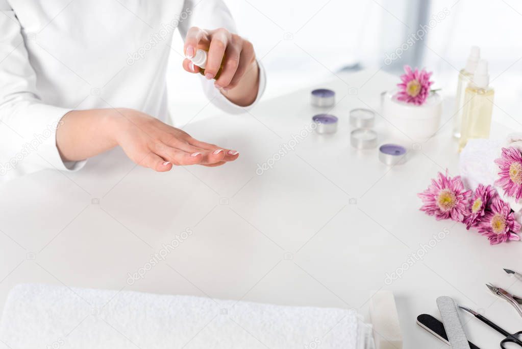 partial view of woman using aroma oil at table with towels, flowers, candles and instruments for manicure in beauty salon