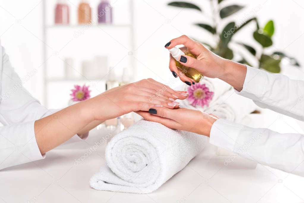 partial view of manicurist spraying aroma oil on hands of woman at table with flowers and towels in beauty salon 