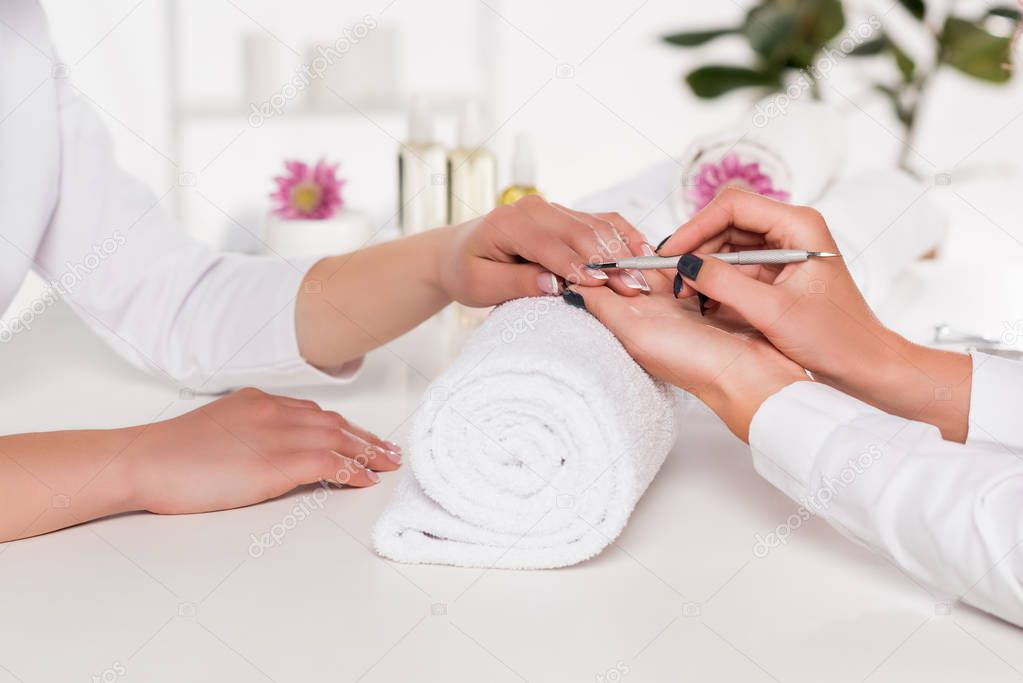 cropped shot of woman receiving manicure by beautician with cuticle pusher at table with flowers and towels in beauty salon 
