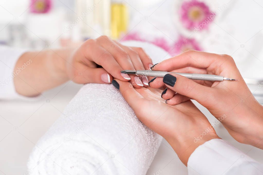 close up image of  woman receiving manicure by beautician with cuticle pusher at table with flowers and towels in beauty salon 