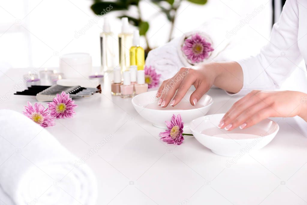 cropped image of woman receiving bath for nails at table with flowers, towels, candles, aroma oil bottles, nail polishes, cream container and tools for manicure in beauty salon 
