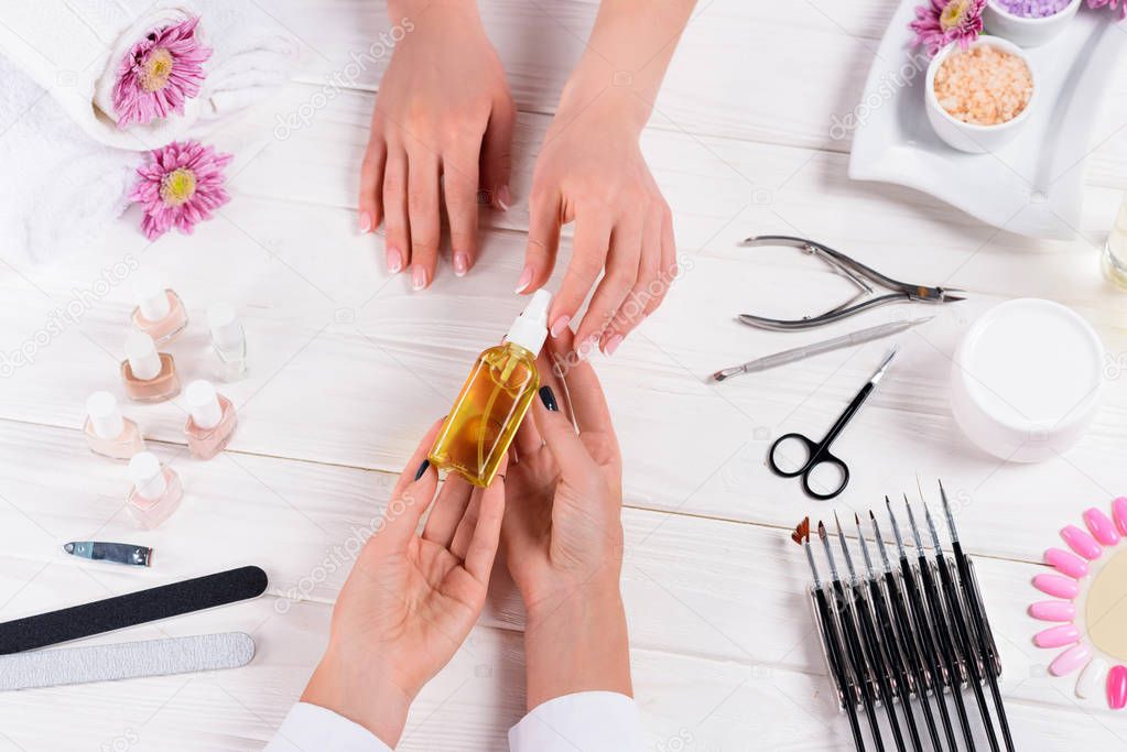 partial view of woman taking aroma oil bottle from hands of manicurist at table with nail polishes, nail files, nail clippers, cuticle pusher, sea salt, flowers, cream and samples of nail varnishes