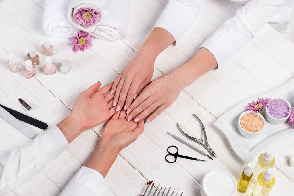 cropped image of manicurist looking at hands of woman at table with flowers, towels, nail polishes, nail files, nail clippers, sea salt, cream, cuticle pusher, scissors and aroma oil bottles