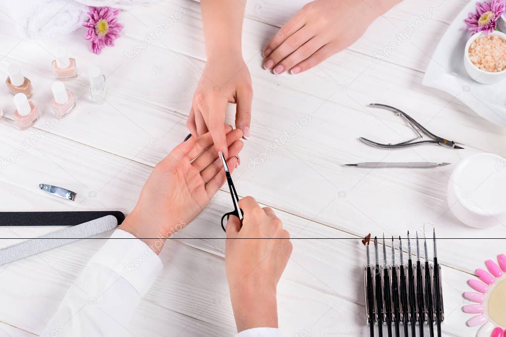 partial view of beautician doing manicure by scissors to woman at table with nail polishes, nail files, nail clippers, cuticle pusher, cream, sea salt, flowers and samples of nail varnishes 