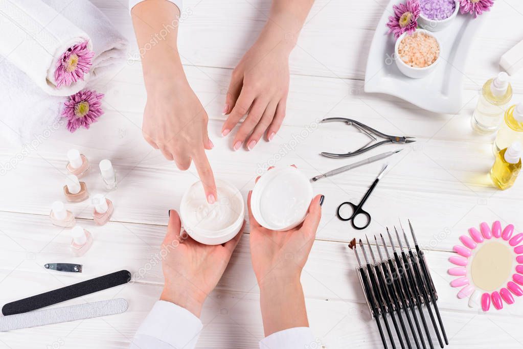 cropped shot of woman pointing on cream in hands of manicurist at table with nail polishes, nail files, nail clippers, cuticle pusher, sea salt, flowers, aroma oil bottles and samples of nail varnishes