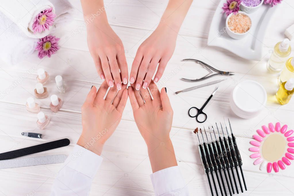 partial view of beautician looking at manicure of woman at table with nail polishes, nail files, nail clippers, cuticle pusher, sea salt, flowers, aroma oil bottles and samples of nail varnishes