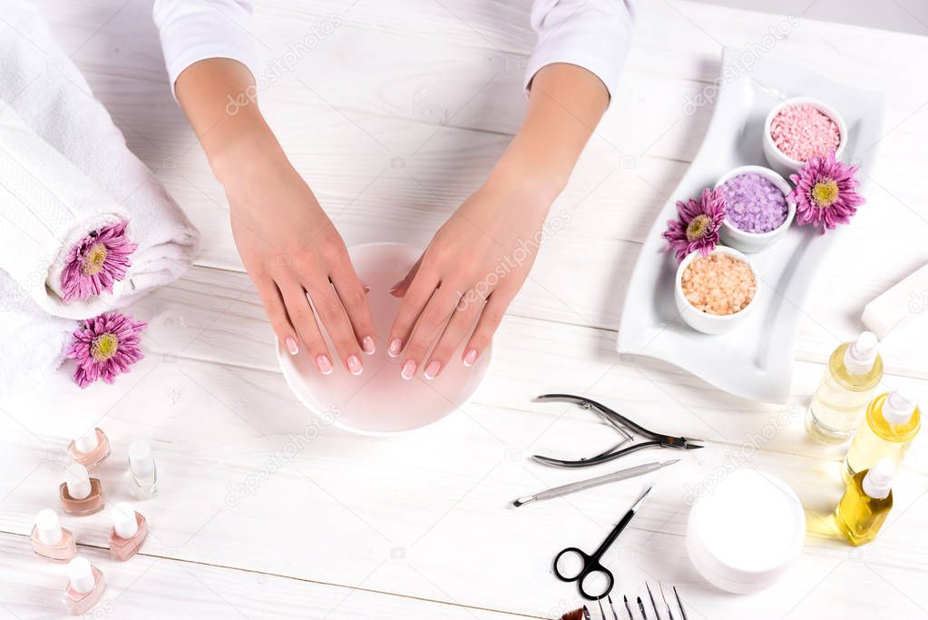 cropped shot of woman receiving bath for nails at table with flowers, towels, colorful sea salt, aroma oil bottles, nail polishes, cream container and tools for manicure in beauty salon 