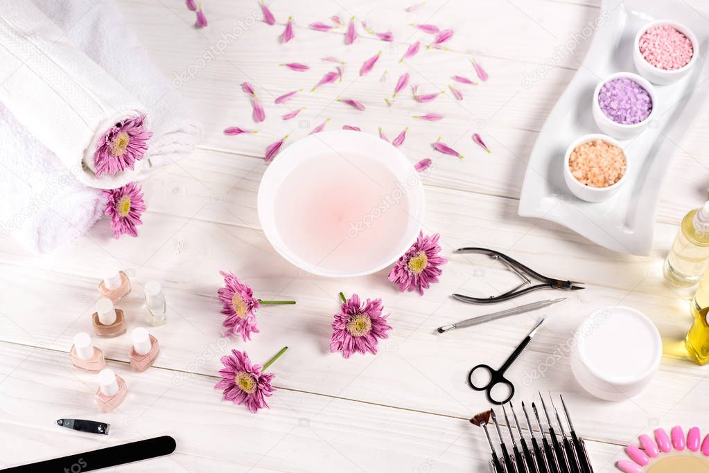 top view of bath for nails at table with flowers, petals, towels, nail polishes, nail file, cuticle pusher, nail clippers, scissors, sea salt, cream container, aroma oil bottles and nail samples of nail varnishes for manicure at table 