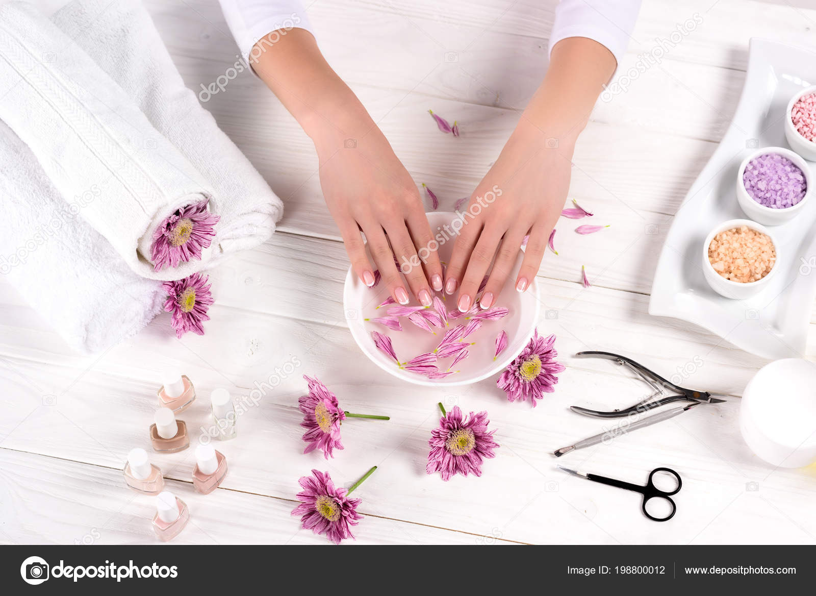 Vista parcial de mulher recebendo banho para unhas na mesa com flores, toalhas, sal do mar colorido, frascos de óleo de aroma, esmaltes, creme recipiente e