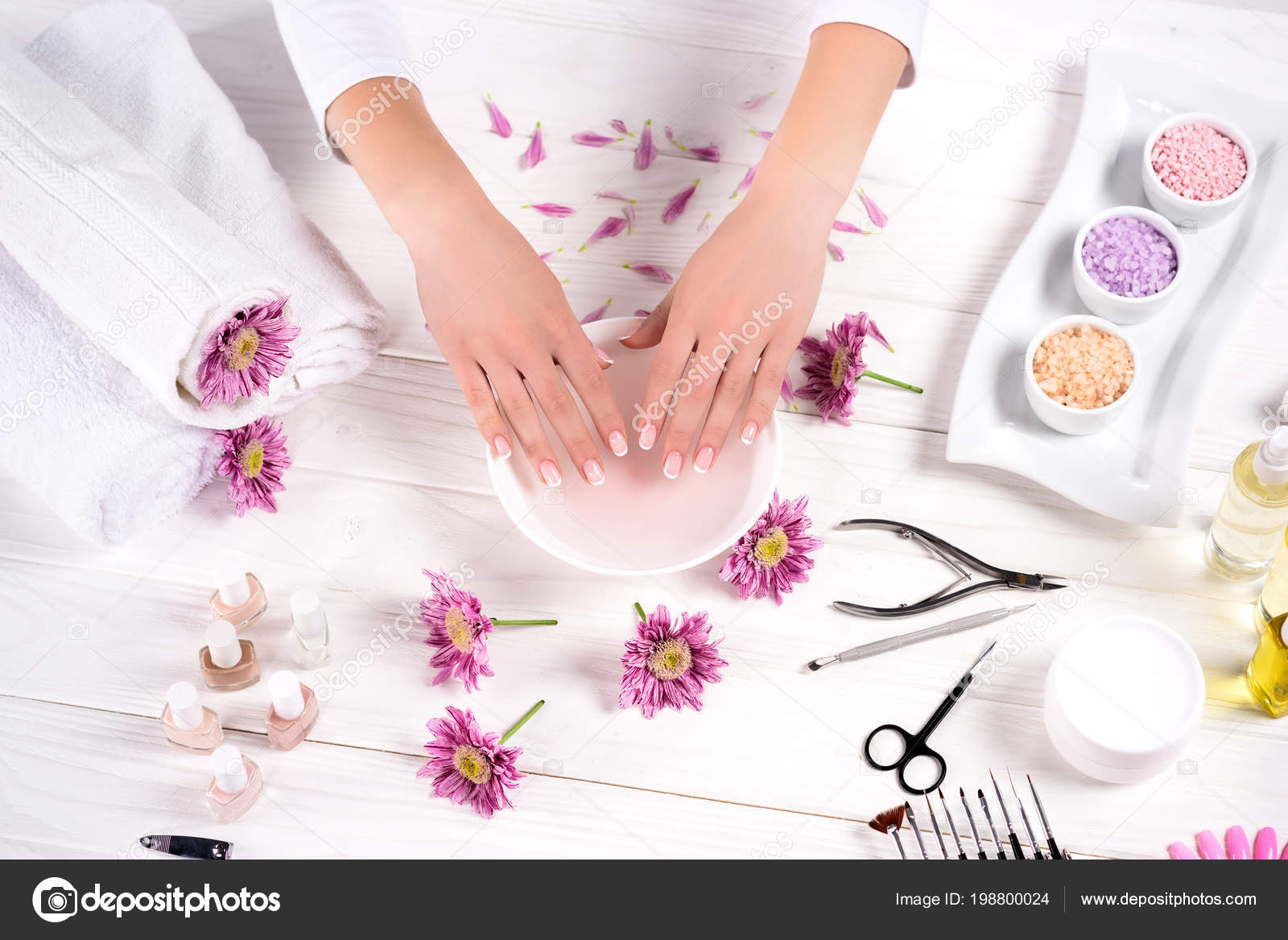 Imagem recortada de mulher recebendo banho para unhas na mesa com flores, toalhas, sal do mar colorido, frascos de óleo de aroma, esmaltes, creme recipiente