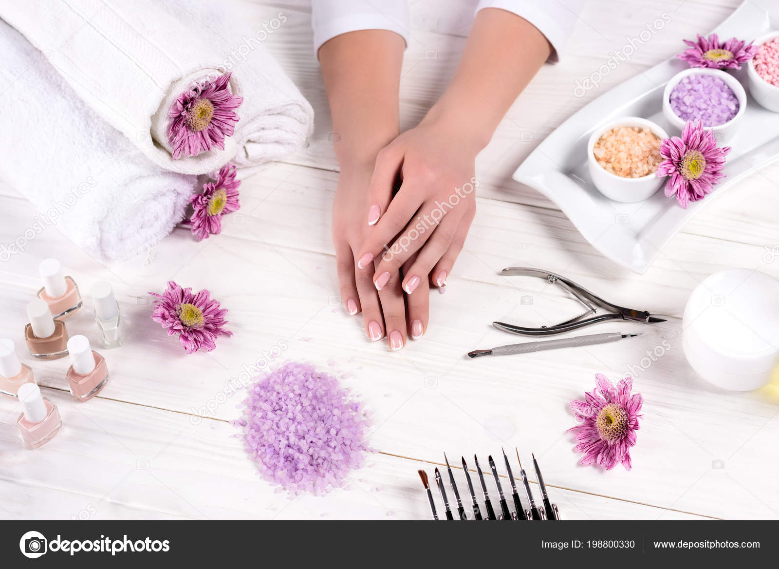 Tiro recortado de mãos femininas na mesa com toalhas, flores, esmaltes, sal do mar colorido, creme recipiente e ferramentas para manicure em salão de beleza