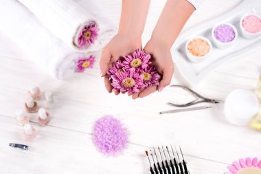 cropped image of woman holding flowers over table with towels, nail polishes, colorful sea salt, cream container and tools for manicure in beauty salon  clipart