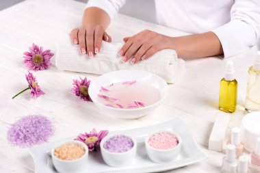 cropped image of woman holding hands on towel for manicure procedure at table with flowers, colorful sea salt, cream container, aroma oil bottles and nail polishes in beauty salon  clipart