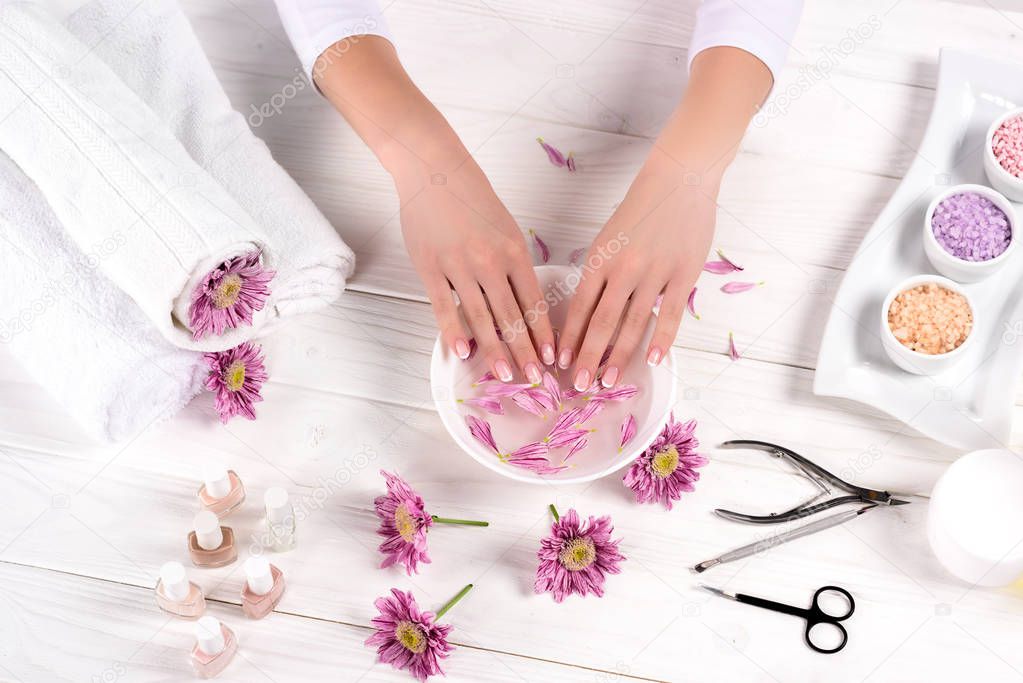 partial view of woman receiving bath for nails at table with flowers, towels, colorful sea salt, aroma oil bottles, nail polishes, cream container and tools for manicure in beauty salon 