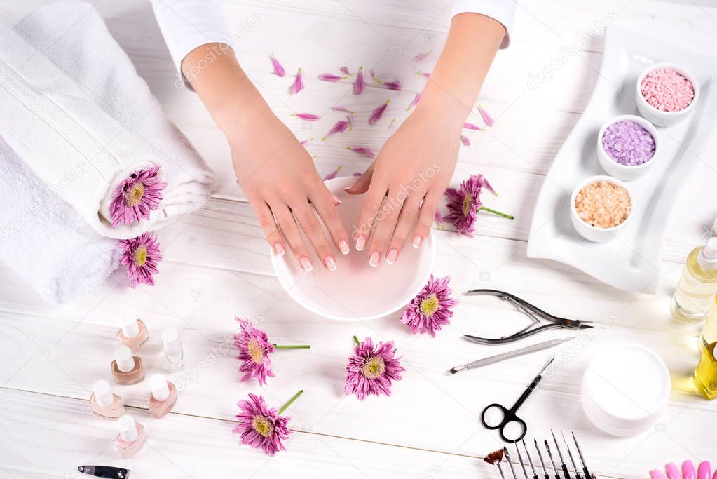 cropped image of woman receiving bath for nails at table with flowers, towels, colorful sea salt, aroma oil bottles, nail polishes, cream container and tools for manicure in beauty salon 